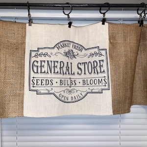 Op de afbeelding: Een jute valance met een centraal paneel met een zwart-wit "GENERAL STORE" bord. Het bord bevat de woorden "MARKET FRESH", "SEEDS", "BULBS", "BLOOMS" en "OPEN DAILY". De valance hangt aan een zwarte stang met metalen ringen.