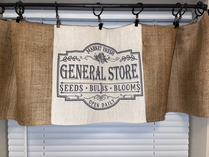 Op de afbeelding: Een jute valance met een centraal paneel met een zwart-wit "GENERAL STORE" bord. Het bord bevat de woorden "MARKET FRESH", "SEEDS", "BULBS", "BLOOMS" en "OPEN DAILY". De valance hangt aan een zwarte stang met metalen ringen.