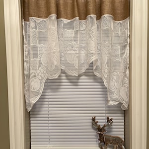 May include: A burlap and lace valance with a white background and a brown burlap top. The valance is hung on a window with white blinds and a wooden reindeer figurine on the window sill.