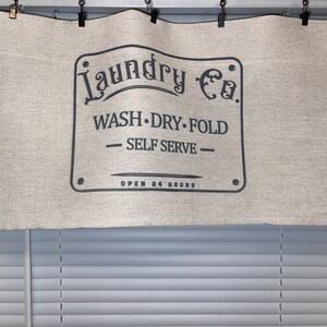 May include: A beige fabric valance with a black sign that reads "Laundry Co. Wash. Dry. Fold - Self Serve - Open 24 Hours".