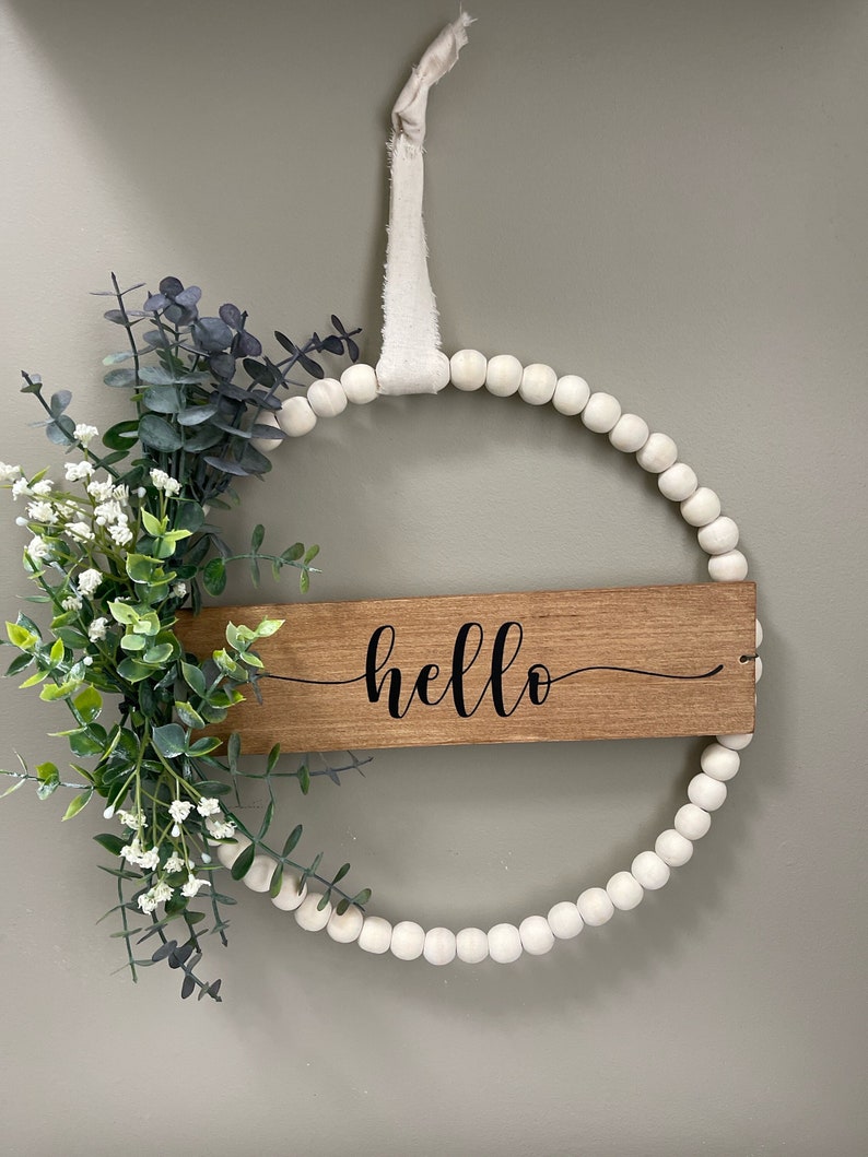 May include: A white bead wreath with a wooden sign that says 'hello' in black cursive lettering. The wreath is decorated with green and blue eucalyptus leaves and white flowers.