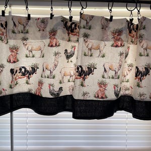 May include: A window valance featuring a repeating pattern of farm animals including cows, sheep, pigs, and roosters, each wearing a small green wreath on its head. The fabric has a cream background and a solid black border along the bottom edge.