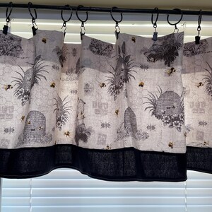 May include: Window curtains featuring a bee and honeycomb design. The curtains have a light background with black and grey illustrations of bees, beehives, and plants. A dark blue border is at the bottom. The curtains are suspended from black rings.