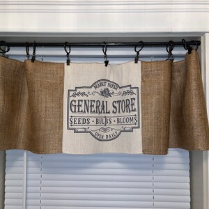 Op de afbeelding: Juten raamgordijn met een centraal paneel met de tekst "GENERAL STORE" in een vintage ontwerp. De raamgordijn is opgehangen aan een zwarte stang boven een raam met witte jaloezie&euml;n. De tekst bevat ook "SEEDS BULBS BLOOMS OPEN DAILY".