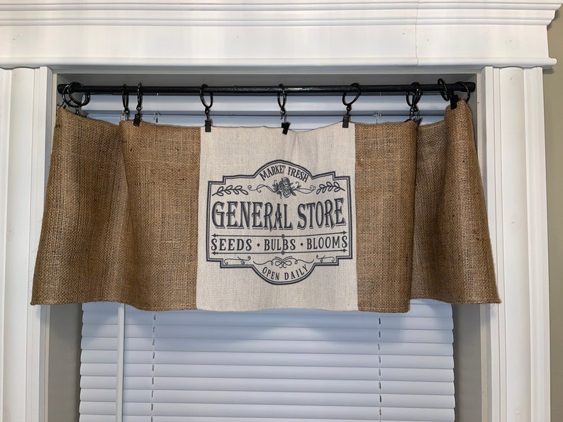 Op de afbeelding: Juten raamgordijn met een centraal paneel met de tekst "GENERAL STORE" in een vintage ontwerp. De raamgordijn is opgehangen aan een zwarte stang boven een raam met witte jaloezie&euml;n. De tekst bevat ook "SEEDS BULBS BLOOMS OPEN DAILY".
