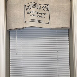 May include: A beige linen valance with black text that reads "Laundry Co. Wash - Dry - Fold - Self Serve - Open 24 Hours" hanging on a black curtain rod above a white window blind.