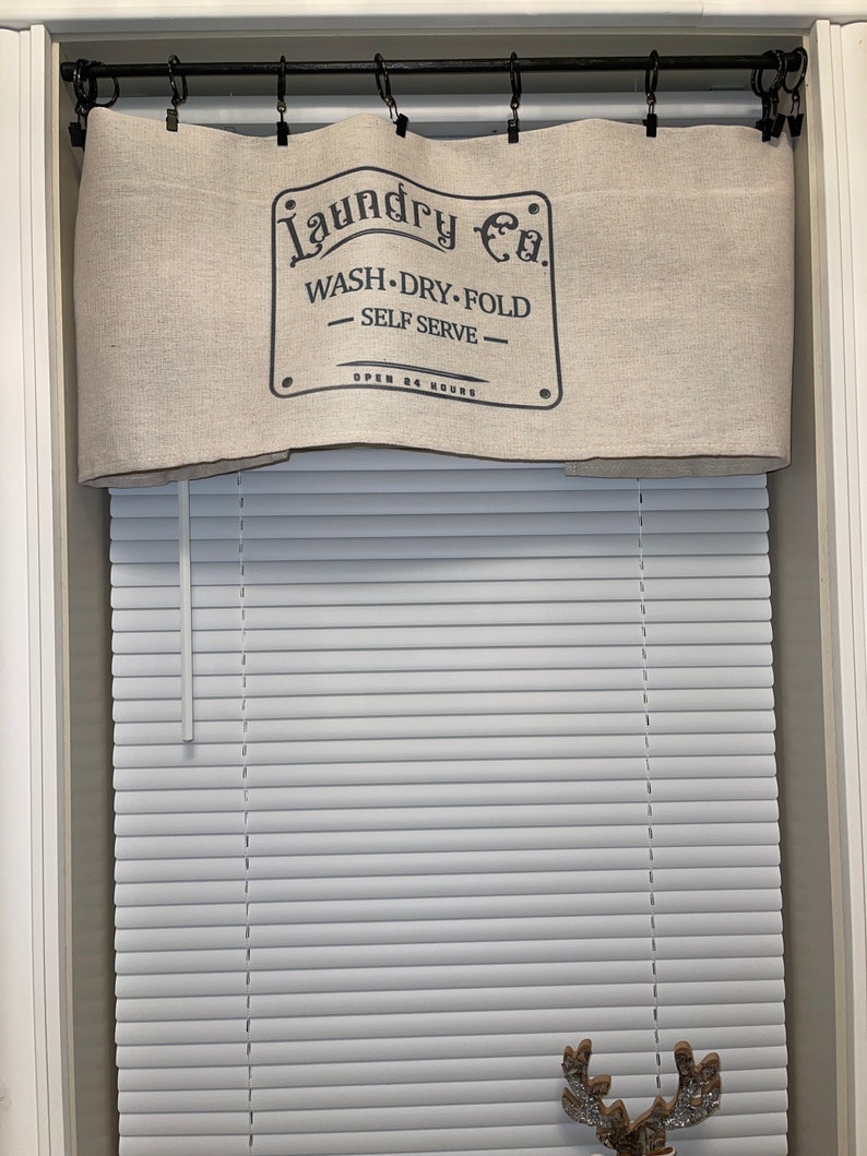 May include: A beige linen valance with black text that reads "Laundry Co. Wash - Dry - Fold - Self Serve - Open 24 Hours" hanging on a black curtain rod above a white window blind.
