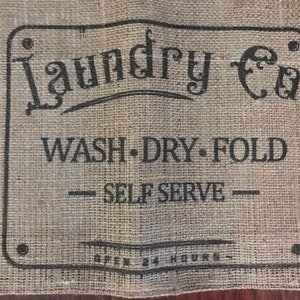 May include: A rectangular burlap sign with the words "Laundry Co." in a decorative font. Below, it reads "WASH • DRY • FOLD - SELF SERVE - OPEN 24 HOURS" in a simple, sans-serif font. The sign has a rustic, vintage aesthetic.