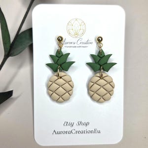 Könnte beinhalten: Handgefertigte Ohrringe in Form von Miniatur-Ananas. Die Ohrringe haben einen beigefarbenen Körper mit einem strukturierten Muster und grünen Blattoberteilen. Sie werden auf einer weißen Karte mit dem Text "Aurora Creation" und "Etsy Shop AuroraCreationEu" präsentiert.
