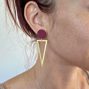 Könnte beinhalten: Ein Hängeohrring mit einem kreisförmigen, texturierten dunkel-kastanienbraunen Stecker, der an einem großen, offenen, goldfarbenen Dreiecksanhänger befestigt ist. Das geometrische Design hängt vertikal am Ohrläppchen und zeigt einen Kontrast zwischen dem matten runden Oberteil und der metallischen dreieckigen Basis.