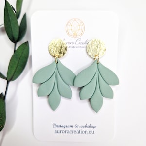 Peut inclure: Une paire de boucles d'oreilles en forme de feuille vert sauge avec une finition mate, suspendues à un clou circulaire doré texturé. Les boucles d'oreilles sont présentées sur une carte blanche avec le texte Aurora Creation et auroracreation.eu.