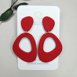 May include: A pair of bright red dangle earrings featuring a textured surface. Each earring consists of a small solid top piece connected to a larger open-center organic shape by a gold-toned metal ring. The earrings are displayed on a white card.