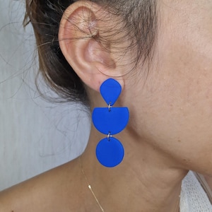May include: Royal blue geometric dangle earrings. The earrings feature a top teardrop shape, a half-circle in the middle, and a full circle at the bottom. The earrings are connected by small metal hoops.