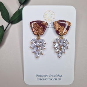 Könnte beinhalten: Ein Paar goldene und burgunderrote Ohrringe mit geometrischer Form und einem Cluster aus klaren Strasssteinen, die an jedem Ohrring hängen. Die Ohrringe werden auf einer weißen Karte mit dem Text "Instagram & webshop auroracreation.eu" präsentiert.