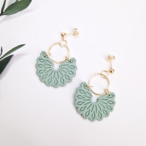 Può includere: Un paio di orecchini verde salvia con un design floreale. Ogni orecchino presenta un pendente a forma di ventaglio smerlato con dettagli incisi, collegato a un cerchio ritorto color oro e un perno dorato. Gli orecchini sono esposti su una superficie bianca.