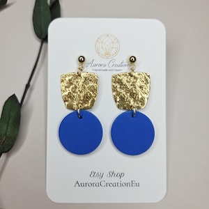 Peut inclure: Une paire de boucles d'oreilles carrées texturées dorées avec un pendentif circulaire bleu. Les boucles d'oreilles sont présentées sur une carte blanche avec le texte "Aurora Creation" et "Etsy Shop AuroraCreationEu".