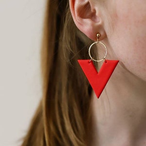 Puede incluir: Primer plano de un pendiente geométrico rojo con un aro y un poste dorados. El pendiente tiene una forma de V audaz. El fondo está borroso, centrándose en el diseño y el color del pendiente.