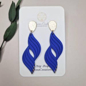 Peut inclure: Une paire de boucles d'oreilles bleues en forme de vagues avec des accents dorés. Les boucles d'oreilles sont présentées sur une carte blanche avec le texte "Aurora Creations" et "Etsy Shop AuroraCreation". Les boucles d'oreilles ont un design texturé.