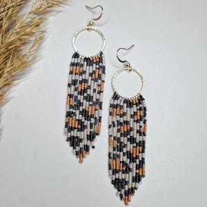 May include: A pair of long, beaded dangle earrings with a gold hoop at the top. The beads are black, white, and gold and are arranged in a leopard print pattern.