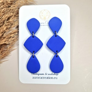 Könnte beinhalten: Ein Paar blaue Ohrringe aus Polymer Clay mit drei geometrischen Formen: einer Träne, einem Diamanten und einem abgerundeten Dreieck. Die Ohrringe werden auf einer weißen Karte mit dem Text "Instagram & webshop aurora creation.eu" gezeigt.