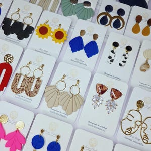 May include: A selection of handmade earrings on display. The earrings showcase diverse designs, including floral, geometric, and abstract shapes, in colours such as blue, yellow, and pink. Some earrings have gold accents and hoops.