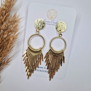 May include: Gold-toned earrings with a textured circular top and a dangling fringe design. The earrings are attached to a white card with the text "Aurora Creations" and a logo. The earrings are displayed on a white surface.