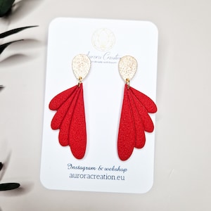 Könnte beinhalten: Ein Paar rote, fächerförmige Ohrringe mit strukturierter Oberfläche, die an goldfarbenen, ovalen Ohrsteckern hängen. Die Ohrringe werden auf einer weißen Karte mit dem Text "auroracreation.eu" präsentiert.
