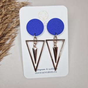 May include: A pair of blue and gold earrings. The earrings feature a round blue disc with a gold triangle pendant. The triangle pendant has a smaller triangle inside. The earrings are on a white card with the text "Instagram & website aurora creation.eu".