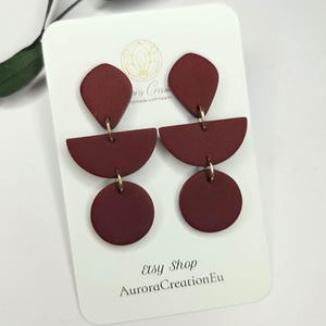 Puede incluir: Pendientes geométricos de color burdeos. Cada pendiente presenta una forma de lágrima, un semicírculo y un círculo, conectados por metal dorado. La tarjeta detrás de los pendientes dice "Etsy Shop AuroraCreationEu".