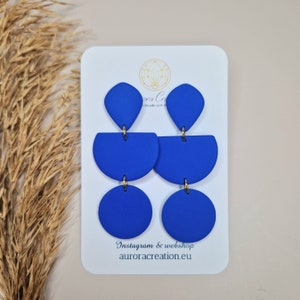 Puede incluir: Un par de pendientes geométricos azules con detalles dorados. Los pendientes están hechos de arcilla polimérica y presentan una forma de lágrima, semicírculo y círculo. Los pendientes se muestran en una tarjeta blanca con el texto "Instagram & webshop aurora creation.eu".