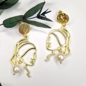 May include: Gold earrings with a pearl dangle and a line drawing of a woman's face. The earrings have a textured disc at the top.