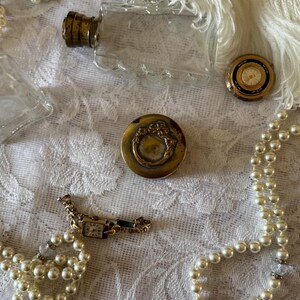 May include: A collection of antique jewellery and accessories, featuring a clear glass perfume bottle with a gold-coloured cap, a small gold-toned compact, a gold-toned watch, and a pearl necklace. The items are displayed on a white lace surface.
