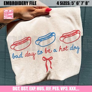 May include: A beige fabric with red and blue embroidered hot dogs and the phrase "bad day to be a hot dog." The design includes a red bow and is labeled as an embroidery file with four size options.
