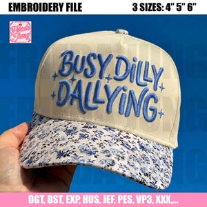 Könnte beinhalten: Cremefarbene Baseballkappe mit dem gestickten Schriftzug "BUSY DILLY DALLYING" in Blau. Der Schirm hat ein Blumenmuster in Weiß und Blau. Erhältlich in 3 Größen: 10,16 cm, 12,7 cm und 15,24 cm.