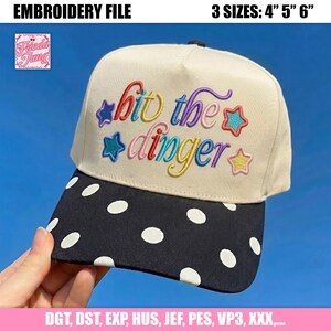 Op de afbeelding: Een crèmekleurige baseballpet met een zwarte klep met witte stippen. Het voorpaneel toont de geborduurde tekst "hit the dinger" in meerkleurige cursieve letters, omringd door kleine geborduurde sterretjes in blauw, rood en geel.