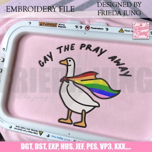Könnte beinhalten: Stickdatei mit einer Gans, die einen Regenbogenumhang trägt und den Text "GAY THE PRAY AWAY". Das Design befindet sich auf einem rosa Stoff, mit "EMBROIDERY FILE" und "DESIGNED BY FRIEDA JUNG" oben. Dateiformate sind unten aufgeführt.