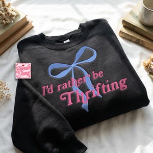 Puede incluir: Sudadera negra con un lazo azul claro y la frase bordada en rosa "I'd rather be Thrifting". La sudadera tiene cuello redondo y mangas largas. Un pequeño logotipo es visible en la esquina.