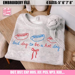 Pu&ograve; includere: Una felpa grigio chiaro con hot dog ricamati rossi e blu e la frase "bad day to be a hot dog". Il design include un fiocco rosso. L'immagine mostra anche "EMBROIDERY FILE" e le opzioni di taglia.
