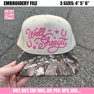 May include: A beige baseball cap with a camouflage print brim. The front features pink embroidered cursive text that reads Well u Sheeyit, accented with small pink stars and horseshoe symbols. The design is presented as an embroidery file.