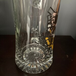 May include: A clear glass beer mug with a handle and a Mickey Mouse design. The design features Mickey Mouse in a yellow and black outfit with the text "Mickey's" in black.