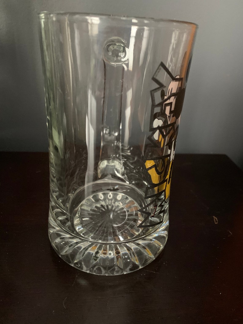 May include: A clear glass beer mug with a handle and a Mickey Mouse design. The design features Mickey Mouse in a yellow and black outfit with the text "Mickey's" in black.