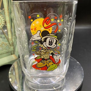 May include: A clear glass with a Mickey Mouse design celebrating his 100th birthday. The glass features Mickey Mouse dressed as an explorer with a red ribbon and confetti. The bottom of the glass is etched with the year 2000.