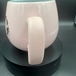 May include: A light pink ceramic mug with a rounded body and a thick handle. The exterior features a black and white line art illustration of a female character with dark hair on the side. The interior of the mug is a pale, light blue colour.