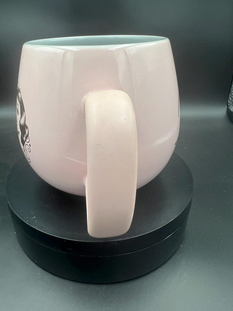 May include: A light pink ceramic mug with a rounded body and a thick handle. The exterior features a black and white line art illustration of a female character with dark hair on the side. The interior of the mug is a pale, light blue colour.