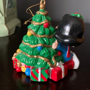 May include: A ceramic Christmas ornament featuring a cartoon character decorating a green Christmas tree with gold tinsel and colorful ornaments. The character is wearing a blue and white outfit and a black hat. The tree is surrounded by wrapped presents.