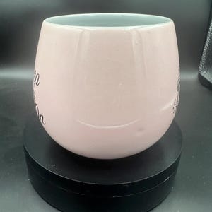 May include: A pale pink ceramic mug with a rounded, bulbous shape and a smooth, glossy finish. The mug features black cursive text printed on the side that reads Keep Going On, with a small decorative graphic element visible on the right side.