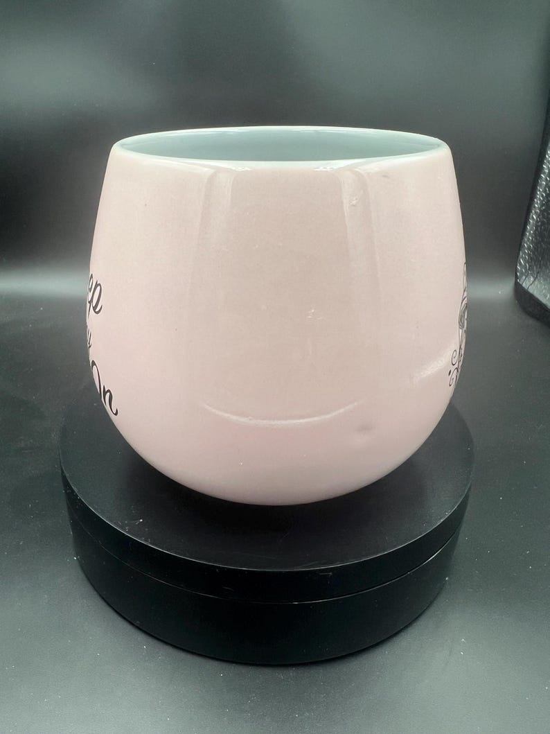 May include: A pale pink ceramic mug with a rounded, bulbous shape and a smooth, glossy finish. The mug features black cursive text printed on the side that reads Keep Going On, with a small decorative graphic element visible on the right side.