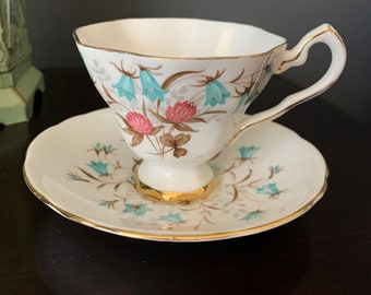 May include: A vintage teacup and saucer set. The white porcelain cup and saucer are decorated with a floral pattern of bluebell flowers and pink clover. Both pieces have gold rims and accents, adding a touch of elegance.