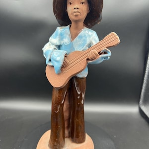 May include: A ceramic figurine of a person playing a guitar. The figure wears a wide-brimmed hat and a light blue shirt with white accents. The guitar is brown, and the trousers are a darker brown. The figurine is on a round black base.