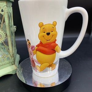 May include: White ceramic mug with a Winnie the Pooh design. Pooh Bear is walking with Piglet and Tigger. The mug has a tall, slender shape and a large handle.
